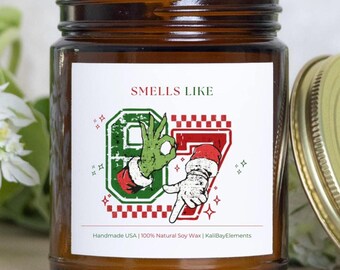 Puede incluir: Una vela en tarro de cristal marrón con una etiqueta blanca. La etiqueta presenta el texto "Smells Like" encima de un gráfico de manos verdes y rojas haciendo el gesto "OK". La etiqueta también incluye el texto "Handmade USA | 100% Natural Soy Wax | KaliBayElements".