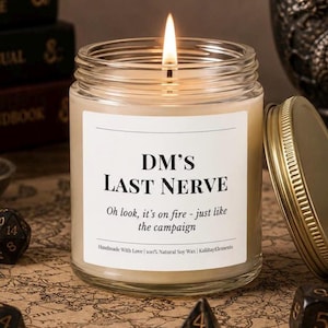 May include: A lit candle in a clear glass jar with a gold lid. The label reads "DM'S LAST NERVE" with the text "Oh look, it's on fire - just like the campaign." The candle is on a map.