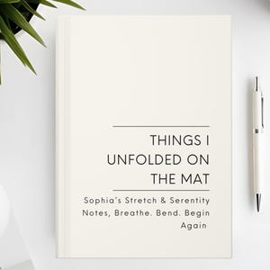 以下が含まれることがあります： 「THINGS I UNFOLDED ON THE MAT」と「Sophia's Stretch & Serenity Notes, Breathe. Bend. Begin Again」の文字が入ったクリーム色のジャーナル。白いペンと植物が白い表面に置かれています。