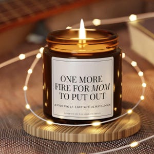 May include: A lit soy wax candle in a brown glass jar with a gold rim, sitting on a wooden coaster. The candle has a white label with the text "ONE MORE FIRE FOR MOM TO PUT OUT". Decorative fairy lights surround the candle.