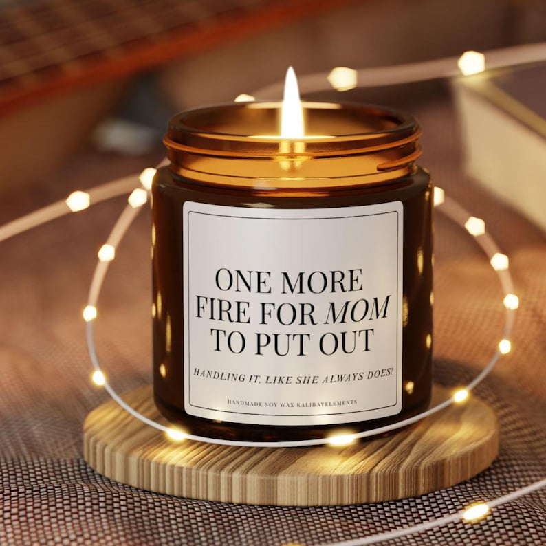 May include: A lit soy wax candle in a brown glass jar with a gold rim, sitting on a wooden coaster. The candle has a white label with the text "ONE MORE FIRE FOR MOM TO PUT OUT". Decorative fairy lights surround the candle.