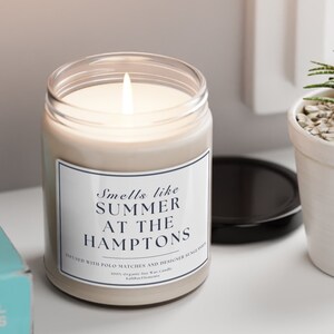 以下が含まれることがあります： 「Smells like Summer at the Hamptons」と「Infused with Polo Matches and Designer Sunglasses」と書かれた黒いラベルが付いた白いキャンドルです。