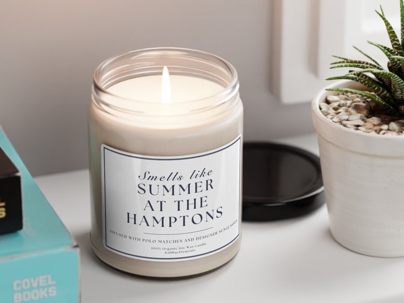 以下が含まれることがあります： 「Smells like Summer at the Hamptons」と「Infused with Polo Matches and Designer Sunglasses」と書かれた黒いラベルが付いた白いキャンドルです。