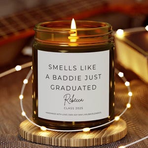 A brown glass candle with a white label that reads "Smells like a baddie just graduated" with the name "Rebecca" and "Class 2025" printed below. The candle is sitting on a wooden coaster.
