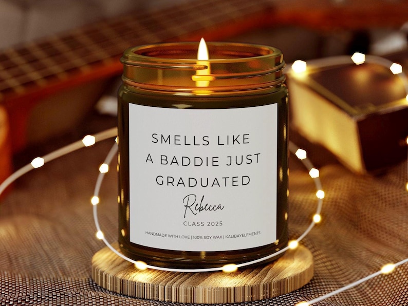 A brown glass candle with a white label that reads "Smells like a baddie just graduated" with the name "Rebecca" and "Class 2025" printed below. The candle is sitting on a wooden coaster.