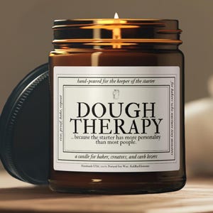 May include: A lit candle in a brown glass jar with a black lid. The label reads "DOUGH THERAPY" and "a candle for baker, creators, and carb lovers." The candle is hand-poured in the USA with natural soy wax.