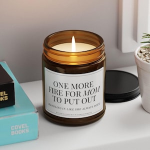 May include: A lit soy wax candle in a brown glass jar with a black lid. The candle has a white label with the text "ONE MORE FIRE FOR MOM TO PUT OUT". The candle is next to a stack of books.