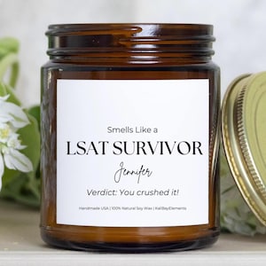 May include: A brown glass jar candle with a white label. The label reads "Smells Like a LSAT SURVIVOR Jennifer Verdict: You crushed it!" The candle has a gold lid and is surrounded by greenery.
