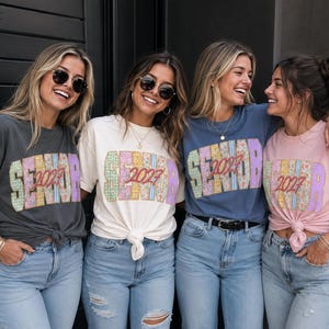 以下が含まれることがあります： チャコール、クリーム、ブルー、ライトピンクの4枚のTシャツ。それぞれに花柄とストライプ模様で「SENIOR」という文字が描かれ、中央には赤い筆記体で「2027」とプリントされています。Tシャツはタックインや結び目を作ったスタイルで、ブルージーンズと合わせています。