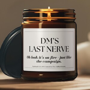 May include: A lit soy wax candle in a brown glass jar with a black lid. The label reads "DM'S LAST NERVE" and "Oh look it's on fire - just like the campaign." Handmade in the USA.