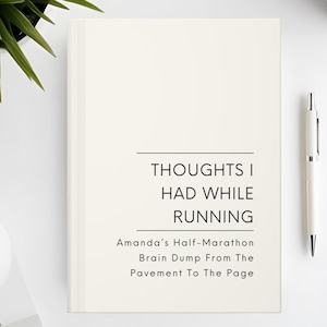 May include: A cream-colored journal with the text "THOUGHTS I HAD WHILE RUNNING" and "Amanda's Half-Marathon Brain Dump From The Pavement To The Page." A white pen and a cup of coffee are on the white surface.