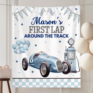 May include: A light blue race car banner with the text "Mason's FIRST LAP AROUND THE TRACK". The banner has a checkered border, blue stars, and a cluster of blue balloons. A vintage gas pump and white balloons are also present.