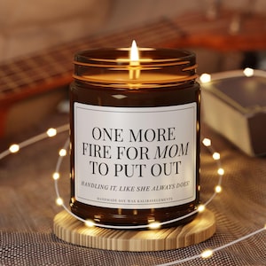 May include: A lit brown glass candle jar with a white label that reads "ONE MORE FIRE FOR MOM TO PUT OUT." The candle sits on a wooden coaster, surrounded by string lights. The text "HANDLING IT, LIKE SHE ALWAYS DOES!" is below the main text.
