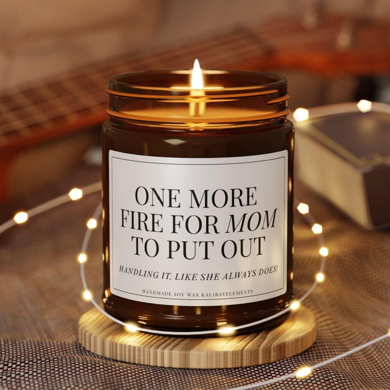 May include: A lit brown glass candle jar with a white label that reads "ONE MORE FIRE FOR MOM TO PUT OUT." The candle sits on a wooden coaster, surrounded by string lights. The text "HANDLING IT, LIKE SHE ALWAYS DOES!" is below the main text.