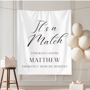 Puede incluir: Bandera blanca con la frase "It's a Match" en elegante escritura negra, felicitando a Matthew, un residente de medicina de urgencias. La escena incluye globos blancos, una caja de regalo y un taburete de madera.