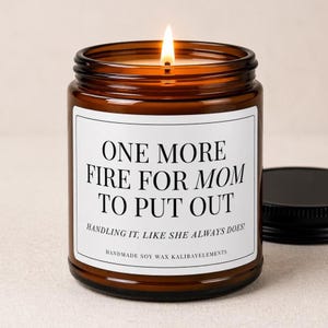 May include: A lit soy wax candle in a brown glass jar with a black lid. The label reads "ONE MORE FIRE FOR MOM TO PUT OUT" with the text "HANDLING IT, LIKE SHE ALWAYS DOES!" and "HANDMADE SOY WAX KALIBAYELEMENTS".