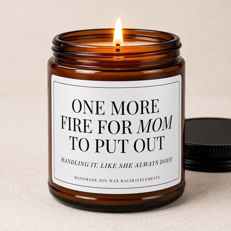 May include: A lit soy wax candle in a brown glass jar with a black lid. The label reads "ONE MORE FIRE FOR MOM TO PUT OUT" with the text "HANDLING IT, LIKE SHE ALWAYS DOES!" and "HANDMADE SOY WAX KALIBAYELEMENTS".