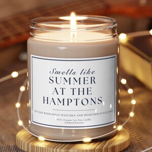 以下が含まれることがあります： 白いラベルに「Smells like SUMMER AT THE HAMPTONS」と「Infused with polo matches and designer sunglasses」と書かれた茶色の大豆ワックスキャンドル。キャンドルに火が灯され、木製の台座の上に置かれています。