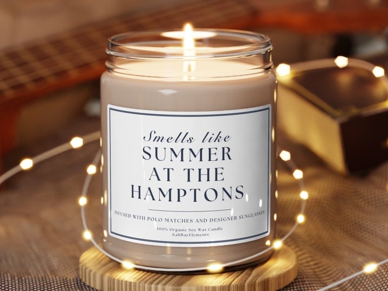 以下が含まれることがあります： 白いラベルに「Smells like SUMMER AT THE HAMPTONS」と「Infused with polo matches and designer sunglasses」と書かれた茶色の大豆ワックスキャンドル。キャンドルに火が灯され、木製の台座の上に置かれています。