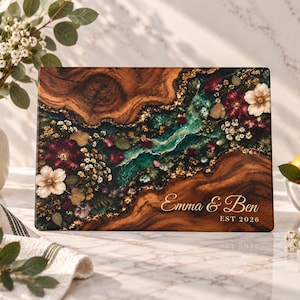 Può includere: Un tagliere rettangolare in legno con un inserto centrale in resina verde scuro, scaglie di foglia d'oro e fiori essiccati. Il tagliere è personalizzato con il testo bianco "Emma & Ben" e "EST 2026" nell'angolo in basso a destra.