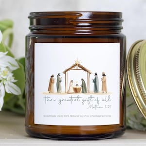 May include: A brown glass jar candle with a white label featuring a Nativity scene illustration. The label includes the text "the greatest gift of all" and "Matthew 1:21". The candle has a gold-colored lid.