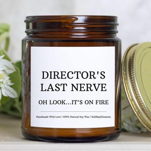 May include: A brown glass candle jar with a white label that reads "DIRECTOR'S LAST NERVE OH LOOK... IT'S ON FIRE" and "Handmade With Love | 100% Natural Soy Wax | KaliBayElements".