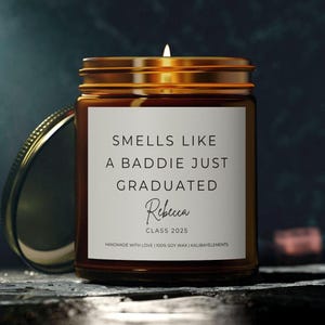 A brown glass candle jar with a gold lid. The label reads "Smells like a baddie just graduated" with the name "Rebecca" and "Class 2025".