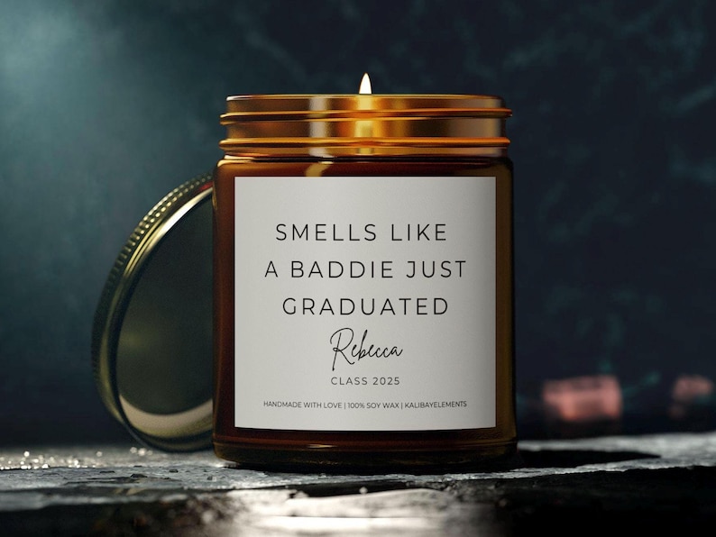 A brown glass candle jar with a gold lid. The label reads "Smells like a baddie just graduated" with the name "Rebecca" and "Class 2025".