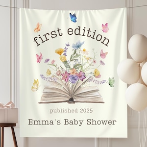 May include: A cream-colored baby shower backdrop with the words "first edition" and "Emma's Baby Shower." The backdrop features an open book with flowers and butterflies. Balloons and a gift box are also visible.