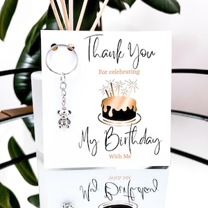 May include: A white card with the text "Thank You For celebrating My Birthday With Me" and a gold and black cake illustration. A silver teddy bear keychain hangs from the top. The card is displayed in front of a plant.