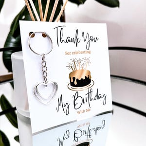May include: A silver heart-shaped keychain hangs from a white card with the text "Thank You For celebrating My Birthday With Me". The card features a gold and black birthday cake illustration.