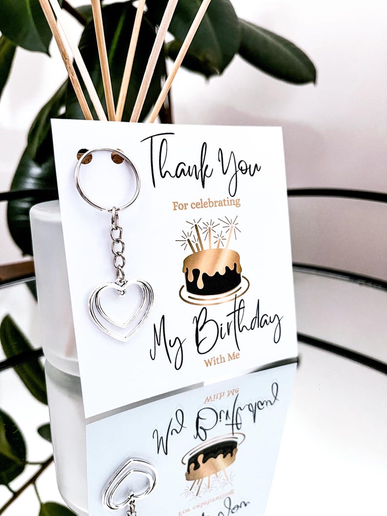 May include: A silver heart-shaped keychain hangs from a white card with the text "Thank You For celebrating My Birthday With Me". The card features a gold and black birthday cake illustration.