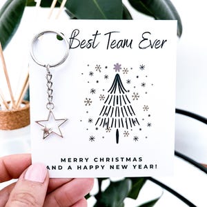 May include: A white card with the text "Best Team Ever" and "MERRY CHRISTMAS AND A HAPPY NEW YEAR!". It features a Christmas tree illustration and a silver star keychain.
