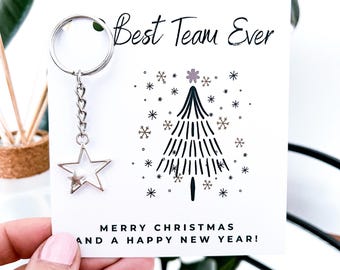 May include: A white card with the text "Best Team Ever" and "MERRY CHRISTMAS AND A HAPPY NEW YEAR!". It features a Christmas tree illustration and a silver star keychain.
