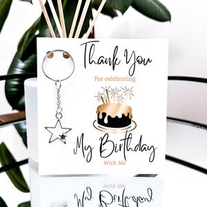 May include: A white card with the text "Thank You For celebrating My Birthday With Me" and a gold and black cake illustration. A silver star keychain hangs from the card. The image is set against a reflective surface.