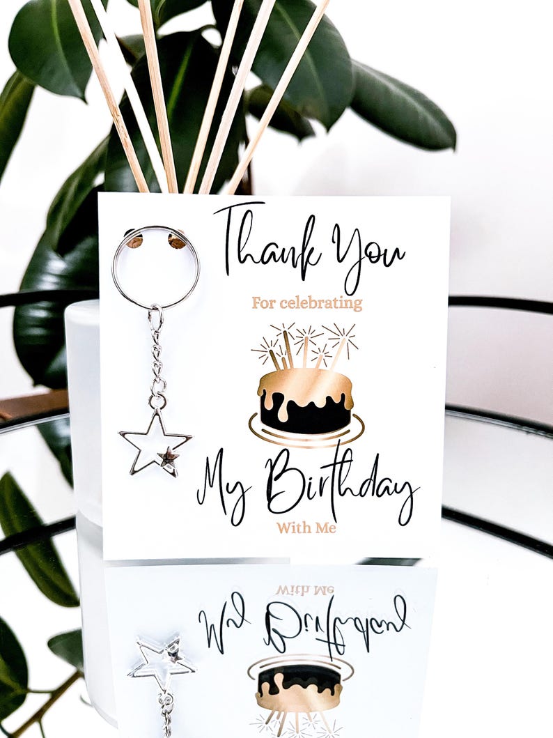 May include: A white card with the text "Thank You For celebrating My Birthday With Me" and a gold and black cake illustration. A silver star keychain hangs from the card. The image is set against a reflective surface.