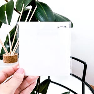 May include: A clear, square, plastic sleeve with a small hole at the top, held in front of a green plant. The sleeve is empty and appears to be designed for holding a card or small item. The background includes a plant and a black metal frame.
