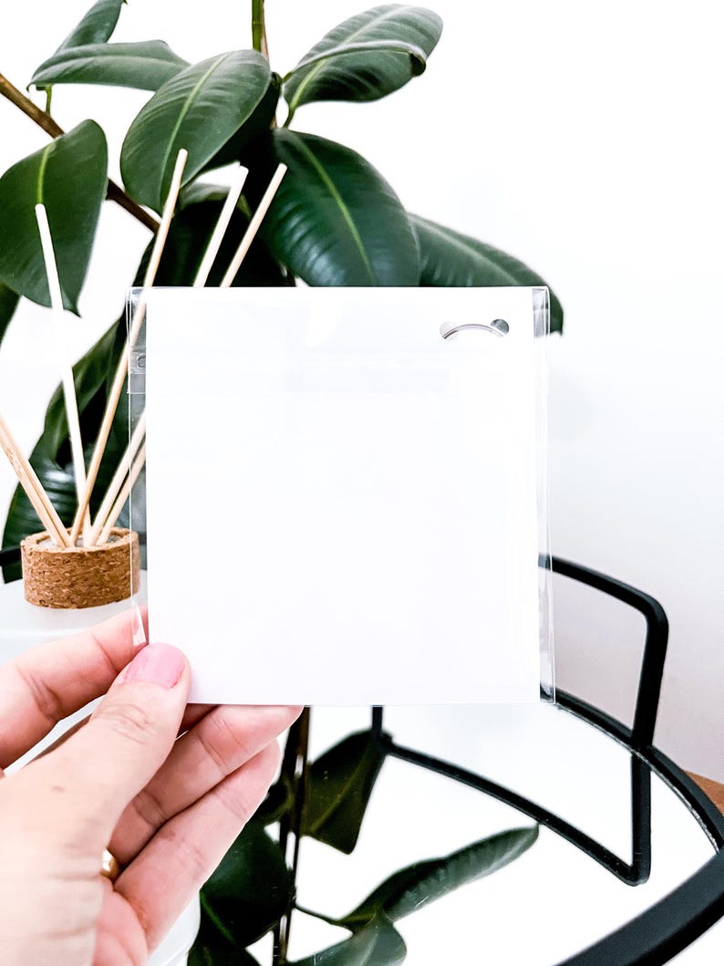 May include: A clear, square, plastic sleeve with a small hole at the top, held in front of a green plant. The sleeve is empty and appears to be designed for holding a card or small item. The background includes a plant and a black metal frame.