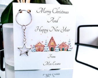 May include: A white card with the text "Merry Christmas and Happy New Year" and "From Mrs Evans" with a silver star charm attached to a silver key ring. The card has a winter scene with gingerbread houses and snow.
