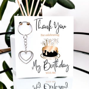 May include: A white card with the text "Thank You For celebrating My Birthday With Me" and a silver heart-shaped keychain. The card features a gold and black cake illustration.