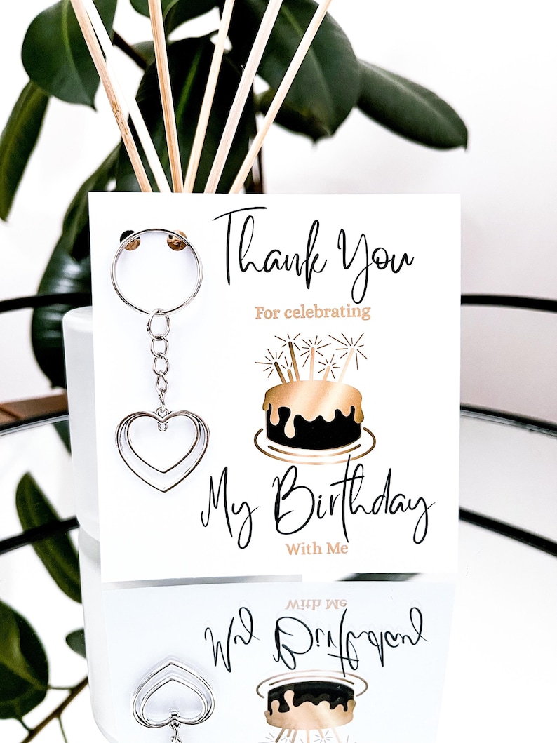May include: A white card with the text "Thank You For celebrating My Birthday With Me" and a silver heart-shaped keychain. The card features a gold and black cake illustration.