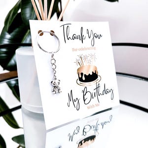 May include: A white card with the text "Thank You For celebrating My Birthday With Me" and a cake illustration. A silver teddy bear keychain hangs from the card. The image is reflected on a mirrored surface.