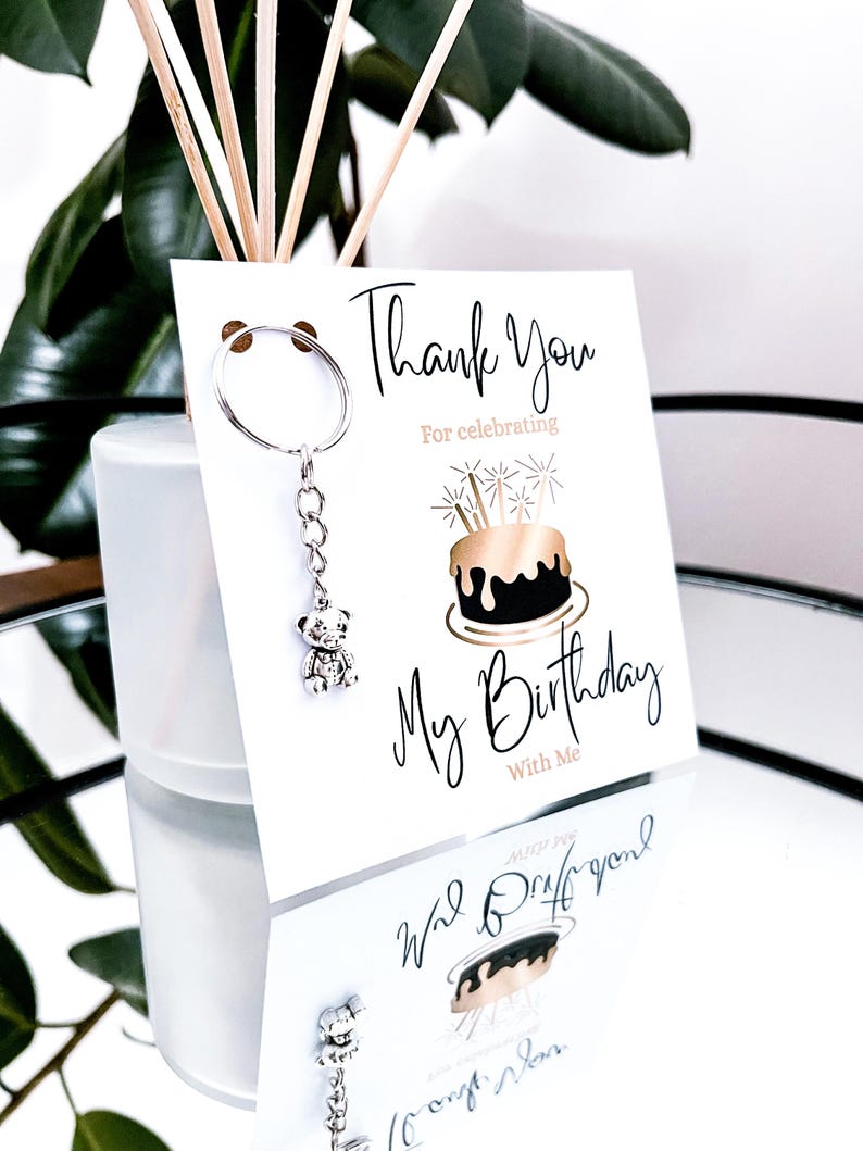 May include: A white card with the text "Thank You For celebrating My Birthday With Me" and a cake illustration. A silver teddy bear keychain hangs from the card. The image is reflected on a mirrored surface.