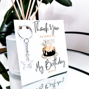May include: A white birthday thank you card with the text "Thank You For celebrating My Birthday With Me". A silver star keychain hangs from the card. The card features a gold and black cake illustration.