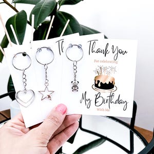May include: A hand holding three white cards with silver keychains. The keychains have heart, star, and teddy bear charms. The card on the right says "Thank You for celebrating My Birthday With Me" with a cake illustration.