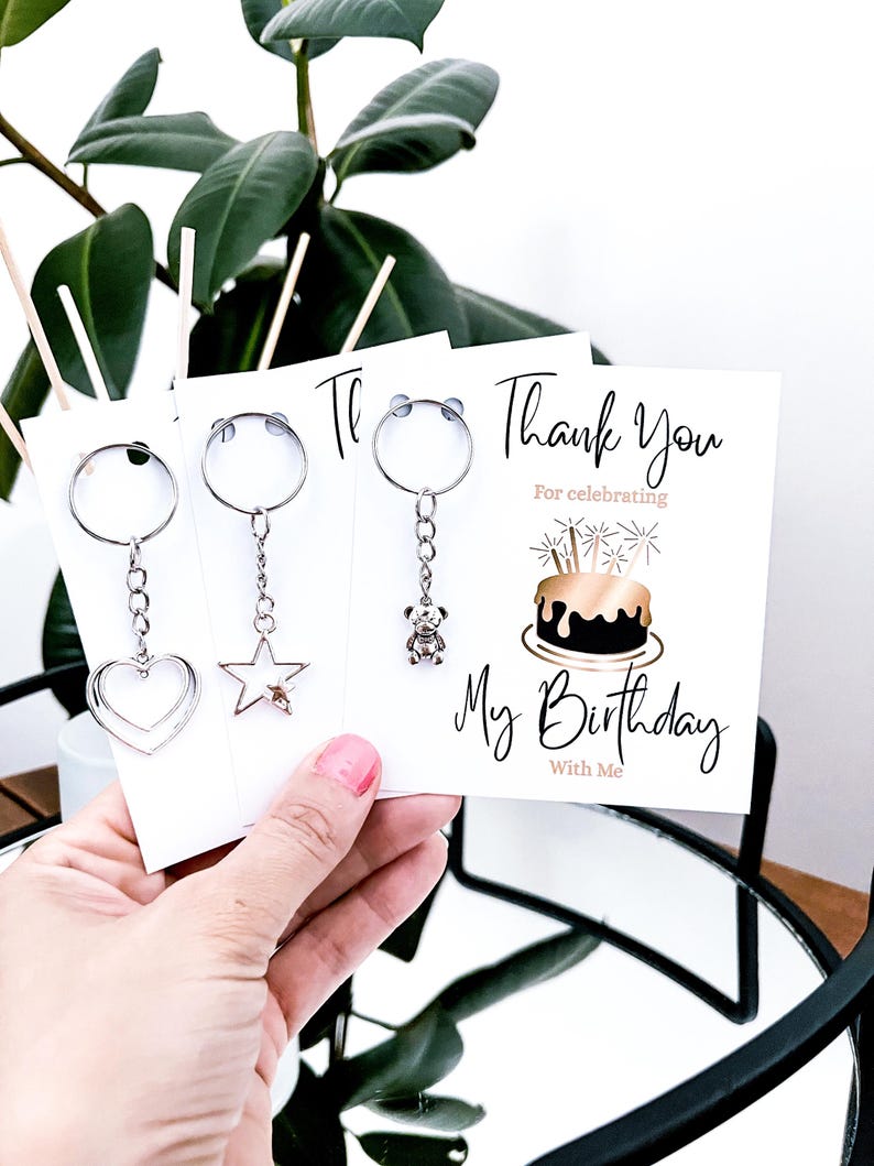 May include: A hand holding three white cards with silver keychains. The keychains have heart, star, and teddy bear charms. The card on the right says "Thank You for celebrating My Birthday With Me" with a cake illustration.