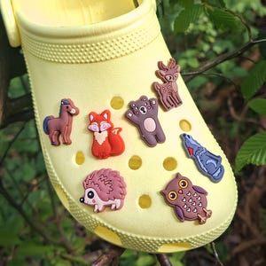 May include: A bright yellow clog shoe decorated with various woodland animal charms. The charms include a brown horse, a red fox, a brown bear, a deer, a blue fish, a hedgehog, and an owl. The shoe is set against a backdrop of green foliage.