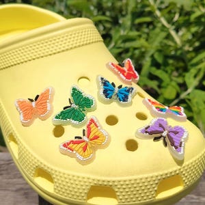 Peut inclure: Un sabot jaune vif orné de breloques papillon colorées. Les papillons sont de différentes couleurs, notamment orange, vert, bleu et violet. La chaussure est en matière caoutchouteuse et a une surface texturée.