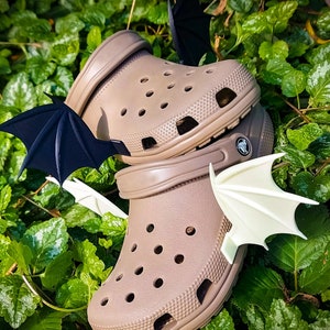 May include: A pair of tan slip-on clogs featuring perforated holes and attached decorative wing accessories. One clog displays black bat-like wings on the side, while the other features white, pointed wing attachments secured to the sides.