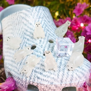 以下が含まれることがあります： 半透明のシロクマのチャームと透明な氷のキューブのチャームで飾られた、青と白の模様のクロッグシューズ。靴は鮮やかなピンクの花に囲まれています。